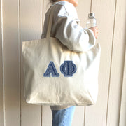 A fashionable college woman poses with her tote bag featuring denim Alpha Phi letters, capturing the spirit of her sorority.