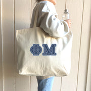 A smiling college woman poses in front of her dorm with a denim Phi Mu tote on her arm.