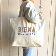 Sigma Delta Tau tote with standout 3D lettering, worn over the shoulder by a woman in Sigma Delta Tau apparel.