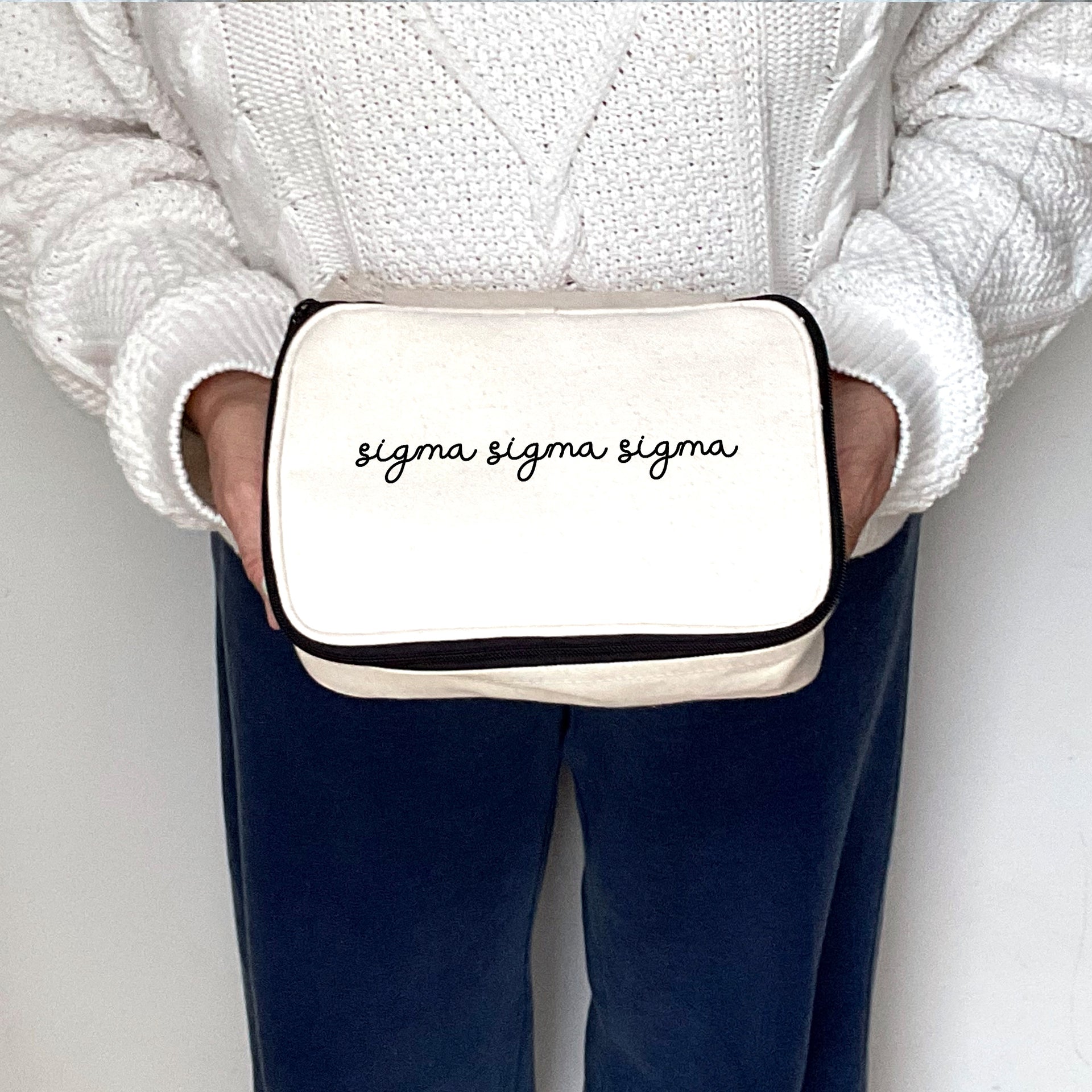 Script name Sigma Sigma Sigma cosmetic pouch held by a casually dressed woman, shown mid-frame.