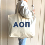 A student in casual campus wear carries her tote with denim Alpha Omicron Pi letters between classes.