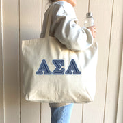 A cheerful student holds her tote bag with denim Alpha Sigma Alpha letters while walking across a sunny quad.