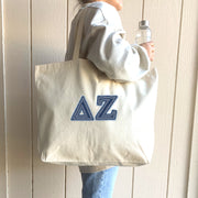 A cheerful woman in a pink hoodie carries a denim Delta Zeta tote while shopping on campus.