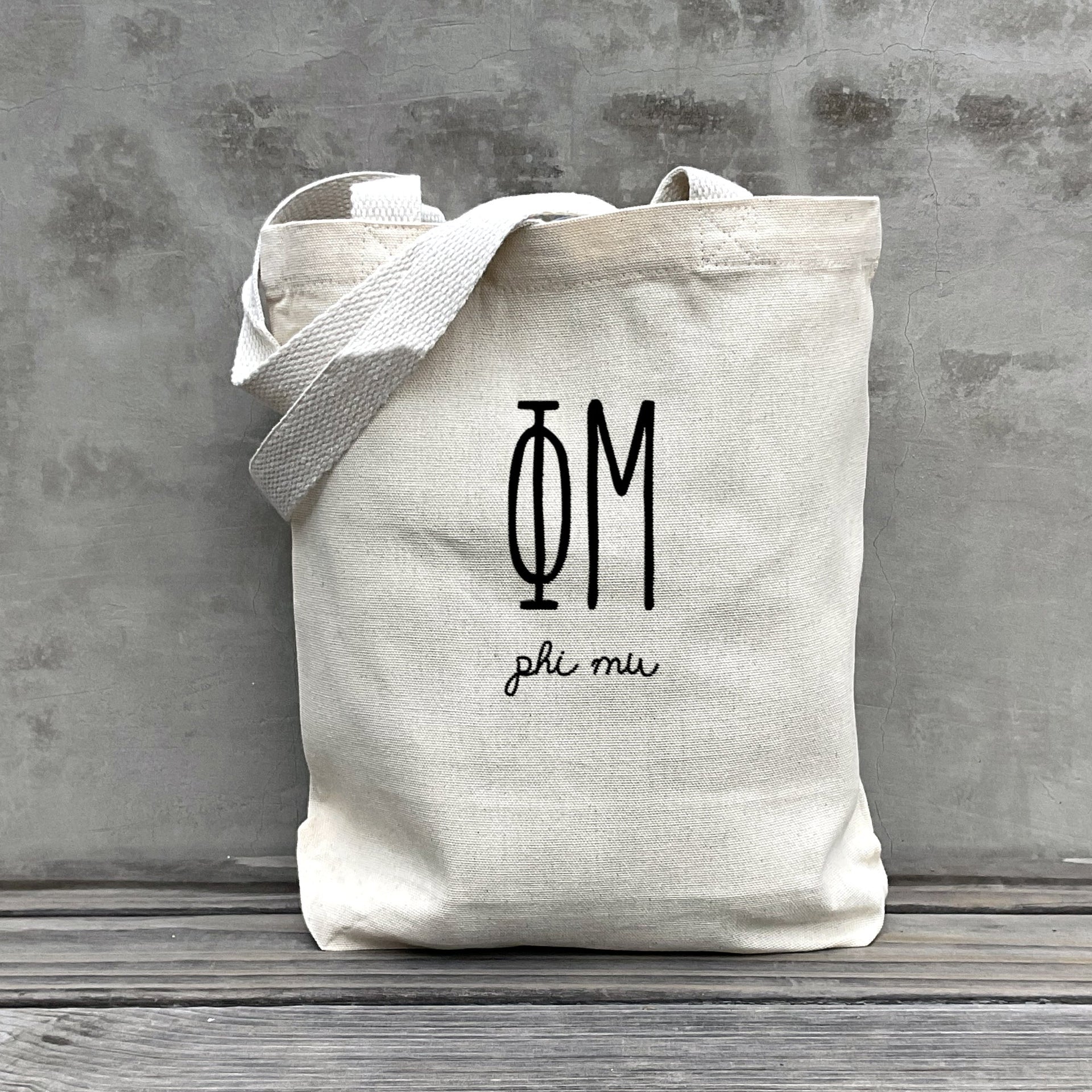 Phi Mu lifestyle image of a Phi Mu tote bag hanging on a dorm door, styled with the sorority’s name in retro font.