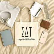 Sigma Delta Tau small canvas tote bag with striped Greek letters styled in a flat lay with everyday essentials